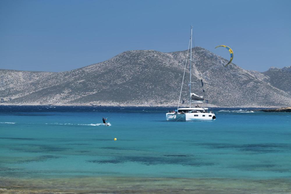 Kitesurfer riding in turquoise waters near a catamaran anchored off a Greek island, surrounded by rugged hills and clear blue skies during a Kite Cruise in Greece.