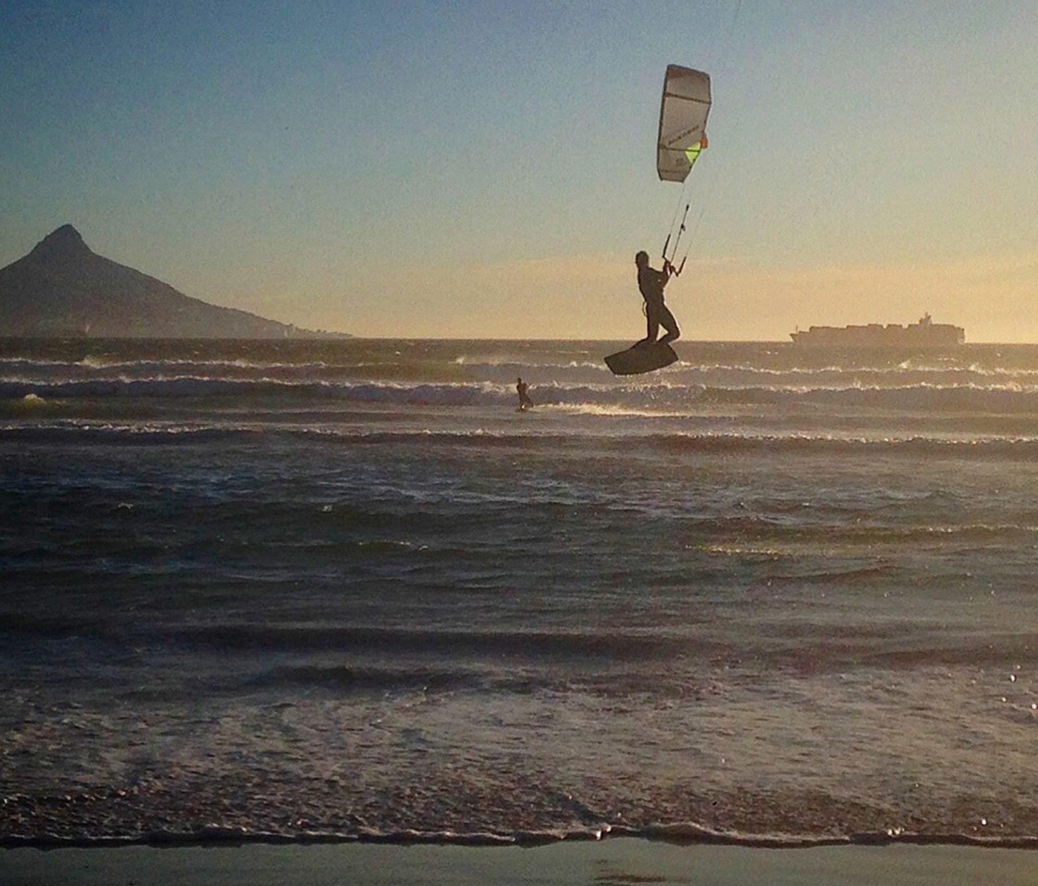 Kitesurfer catching air at sunset near Cape Town, South Africa, with waves breaking along the shore and Lion’s Head mountain visible in the background.
