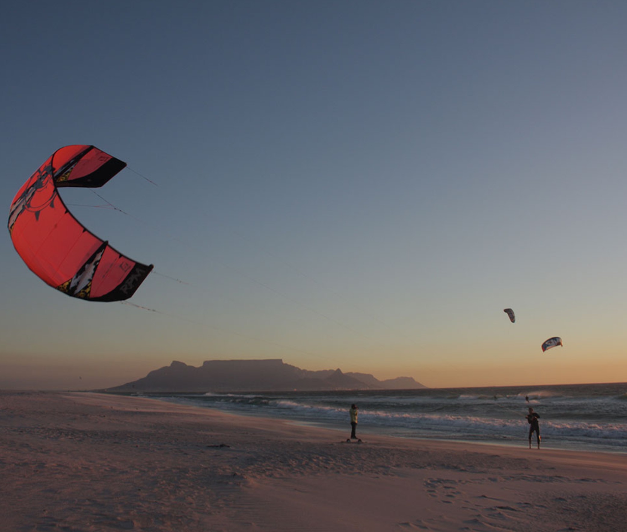 Kitesurfers launching their kites at sunset on a beach near Cape Town, South Africa, with Table Mountain silhouetted in the background against a glowing evening sky.