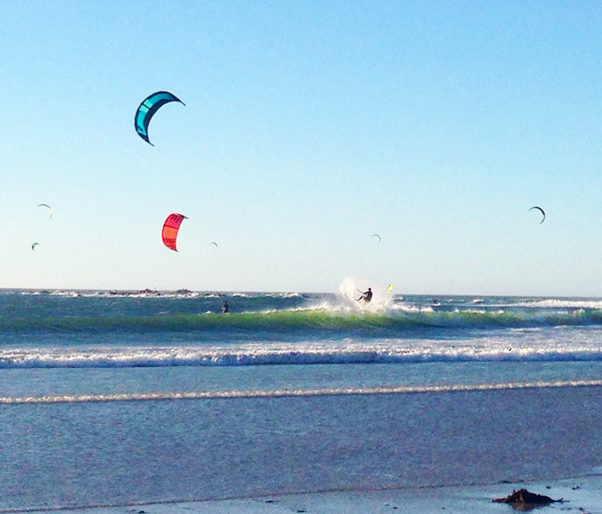 Kitesurfers riding waves at Big Bay near Cape Town, South Africa, with colorful kites flying over the ocean and late-afternoon sunlight illuminating the surf.